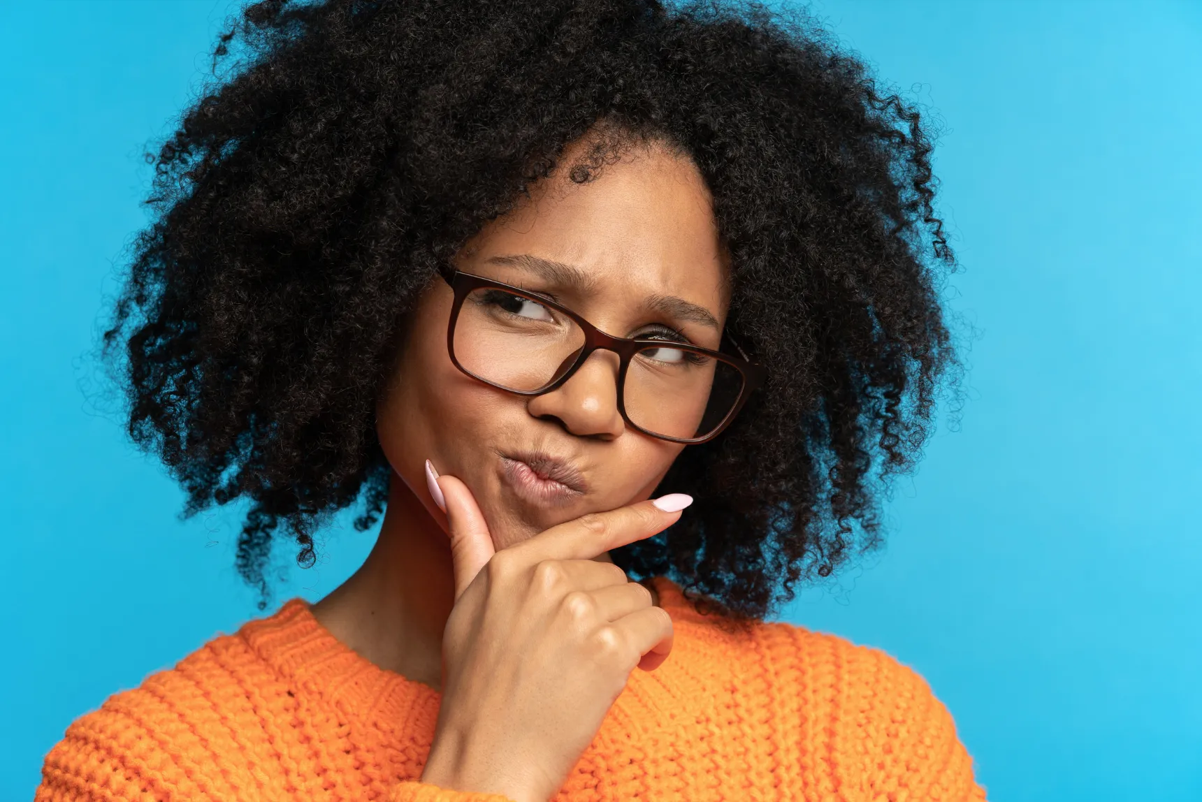 Woman posing with hand on chin. She's wearing glasses and has curly hair, looking towards the camera with a thoughtful expression.