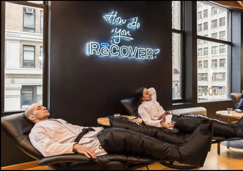 Clients in compression suits reclining beneath the neon How do you RēCOVER? sign