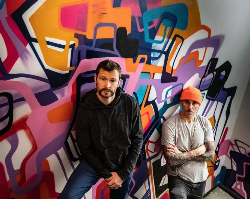 Two men seated in front of a colorful graffiti mural