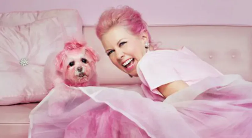 A woman with pink hair lying on a pink bed with a small dog