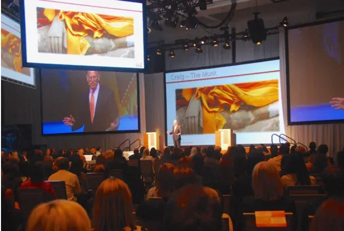 A speaker on stage in front of a large audience and projection screens