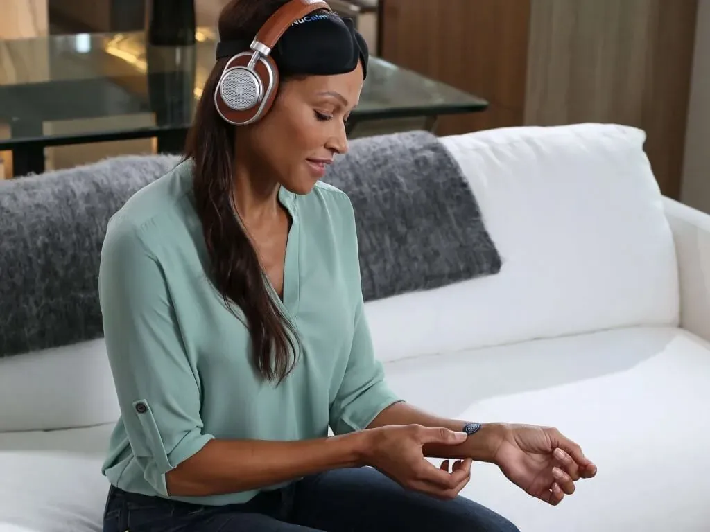 A woman sitting on a sofa wearing NuCalm headphones and an eye mask, applying a biosignal processing disc to her wrist